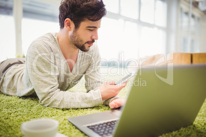 Businessman lying on carpet while using laptop and mobile phone