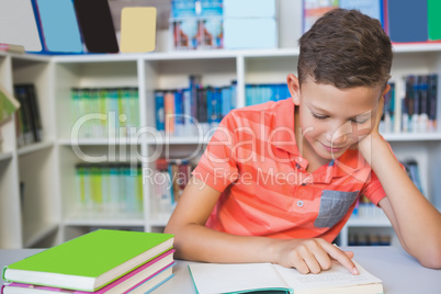 Schoolboy sitting on table and reading book in library