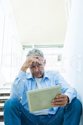 Worried mature man holding tablet while sitting on steps