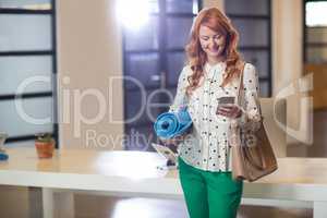 Smiling woman using phone while holding mat in office