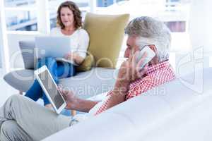Mature man talking on phone while holding tablet at home