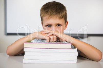 Portrait of sad schoolboy leaning on stack of books in class roo