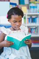 Schoolgirl sitting on chair and reading book in library