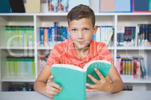 Schoolboy sitting on table and reading book in library