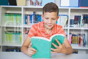 Schoolboy sitting on table and reading book in library