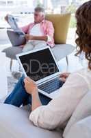 Woman using laptop while mature man reading newspaper