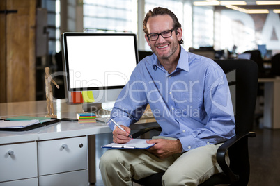 Confident businessman writing on clipboard by desk