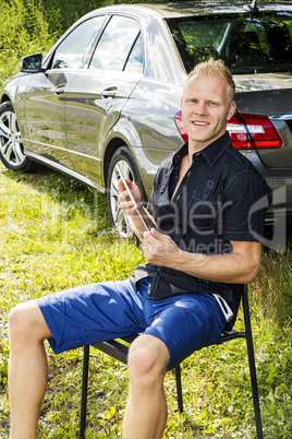 Man sitting with Tablet PC in front of his car in the green