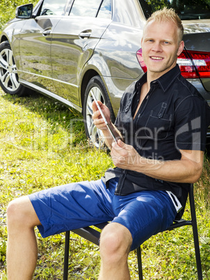 Man sitting with Tablet PC in front of his car in the green