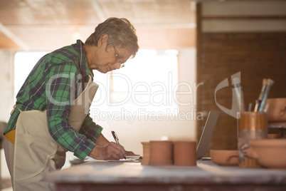 Female potter writing on a book while using laptop