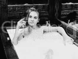 naked young smiling woman with long hair and straight slim beautiful body lying in white bath tub indoor on wooden background. Young woman drinks a champangne. black and white