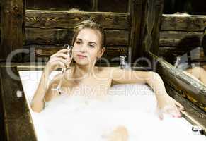 naked young smiling woman with long hair and straight slim beautiful body lying in white bath tub indoor on wooden background. Young woman drinks a champangne. black and white