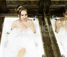 naked young smiling woman with long hair and straight slim beautiful body lying in white bath tub indoor on wooden background. Young woman drinks a champangne. black and white