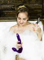 naked young smiling woman lying in white bath tub on wooden background. Young woman drinks a champangne and holding dildo. sex toys