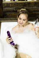 naked young smiling woman lying in white bath tub on wooden background. Young woman drinks a champangne and holding dildo. sex toys