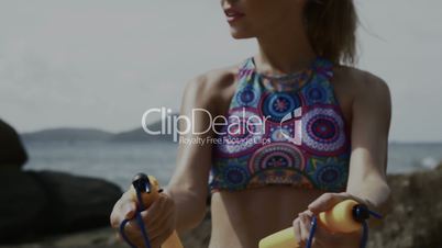 Pretty blonde woman exercising using jumping rope between the rocks near the sea during summer day
