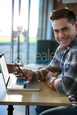 Smiling man using mobile phone with laptop on table