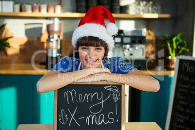 Smiling waitress wearing a santa hat and sitting with a X-mas si
