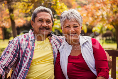 An elderly couple smiling at the camera sitting on the bench