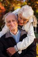 An elderly woman hugs her husband sitting on the bench looking a