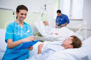 Patient lying on bed while nurse writing on clipboard