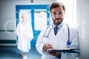 Confident doctor with clipboard standing in hospital corridor