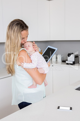 Mother using digital tablet while carrying her baby in kitchen