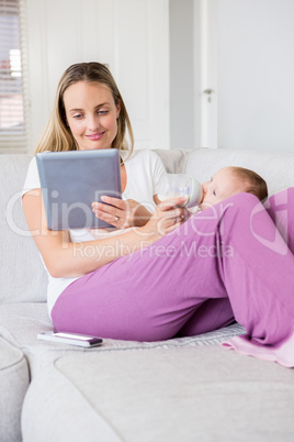 Mother using digital tablet while feeding her baby with milk bottle