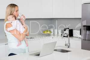 Mother drinking water while carrying baby in kitchen