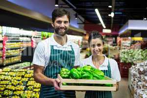 Smiling staff holding a crate of green bell pepper at supermarket