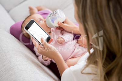 Mother using mobile phone while feeding her baby with milk bottle
