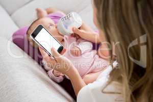 Mother using mobile phone while feeding her baby with milk bottle