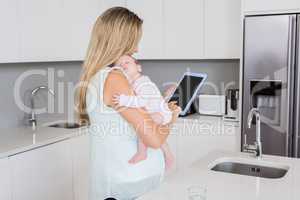 Mother using digital tablet while carrying her baby in kitchen