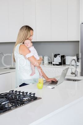 Mother using laptop while carrying baby in kitchen