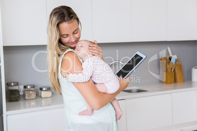 Mother using digital tablet while carrying her baby in kitchen