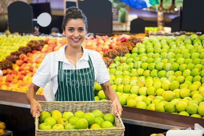 Smiling female staff holding a basket of green apple at supermarket