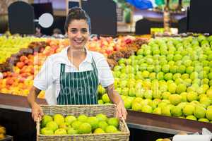Smiling female staff holding a basket of green apple at supermarket