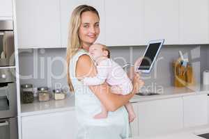 Mother using digital tablet while carrying her baby in kitchen