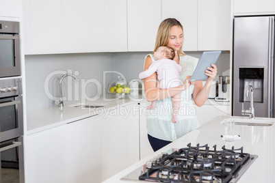 Mother using digital tablet while carrying her baby in kitchen
