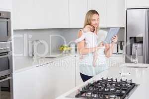 Mother using digital tablet while carrying her baby in kitchen