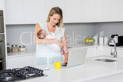 Mother using laptop while carrying baby in kitchen