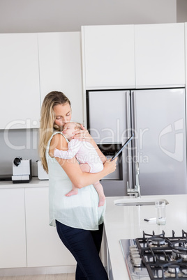 Mother using digital tablet while carrying her baby in kitchen