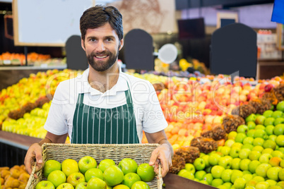 Smiling male staff holding a basket of green apple at supermarket