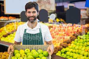 Smiling male staff holding a basket of green apple at supermarket