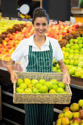 Smiling female staff holding a basket of green apple at supermarket