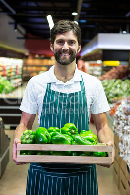 Smiling male staff holding a crate of green bell pepper at supermarket