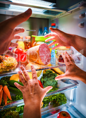 Human hands reaching for food at night in the open refrigerator