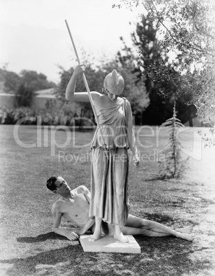 Nude man outside with statue of woman with spear