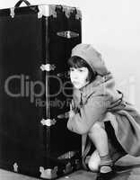 Little girl in hat and coat crouching next to a suitcase
