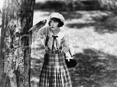 Dressed to kill, a young woman with a knife waits behind a tree